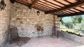 Un patio con paredes de piedra y techo de madera, dos sillas de metal, una mesa pequeña con una cesta de flores, y vistas al exterior con vegetación.