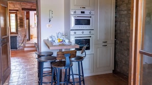 Cocina con alacenas blancas, horno doble y pared de piedra. Hay una mesa con sillas y un frutero.