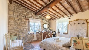 Un dormitorio con pared de piedra, vigas de madera en el techo, una cama con ropa de cama floral, una mesita de noche, un aparador, un espejo y una araña.