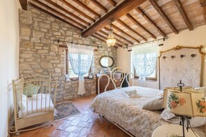 A bedroom with a stone wall, wooden ceiling beams, a bed with floral bedding, a nightstand, a dresser, a mirror, and a chandelier. - NEW - The Lost Stone country paradise with pool, AC & breathtaking country views (Todi - PG)