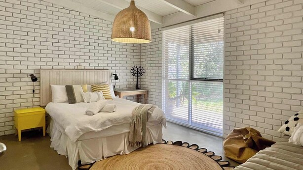 Une chambre avec un mur en briques blanches, un lit avec une literie blanche, un tapis rond en fibres naturelles, un luminaire suspendu et une fenêtre avec des stores.