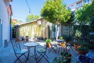 Un patio al aire libre con mesas y sillas redondas, rodeado de plantas en macetas y un árbol.
