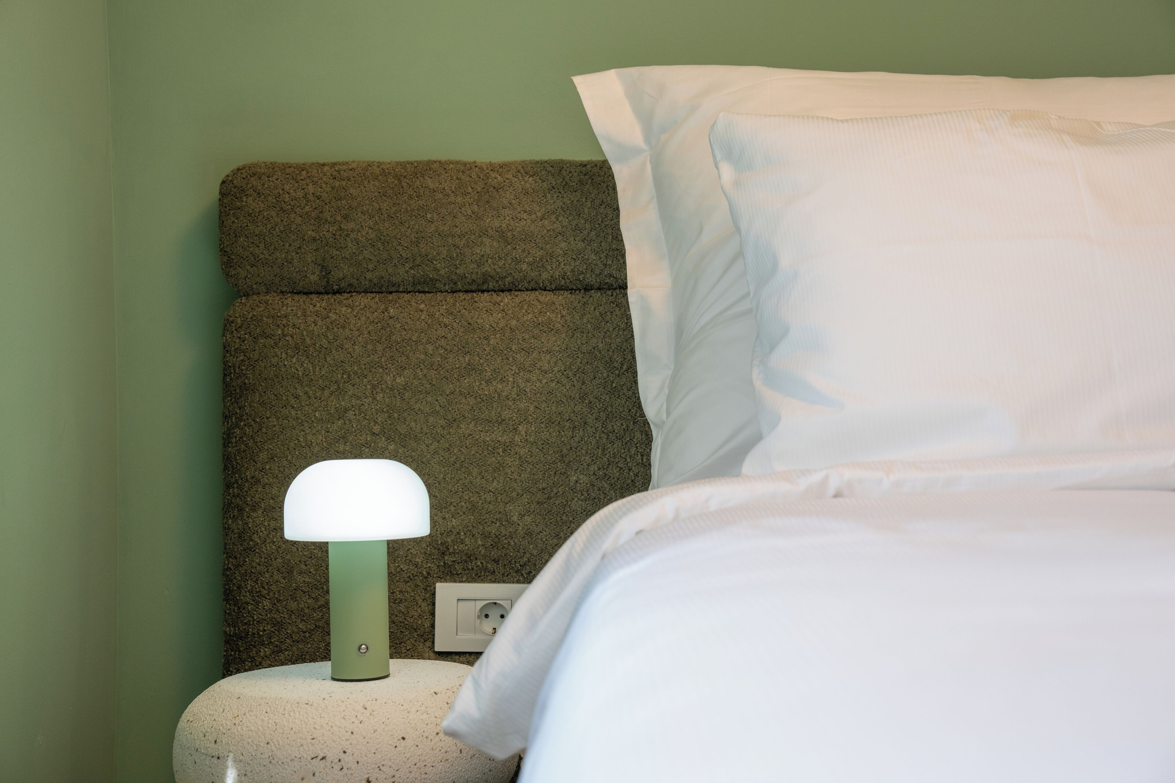 Una cama bien hecha con sábanas blancas, un cabecero verde con textura y una lámpara de noche blanca.