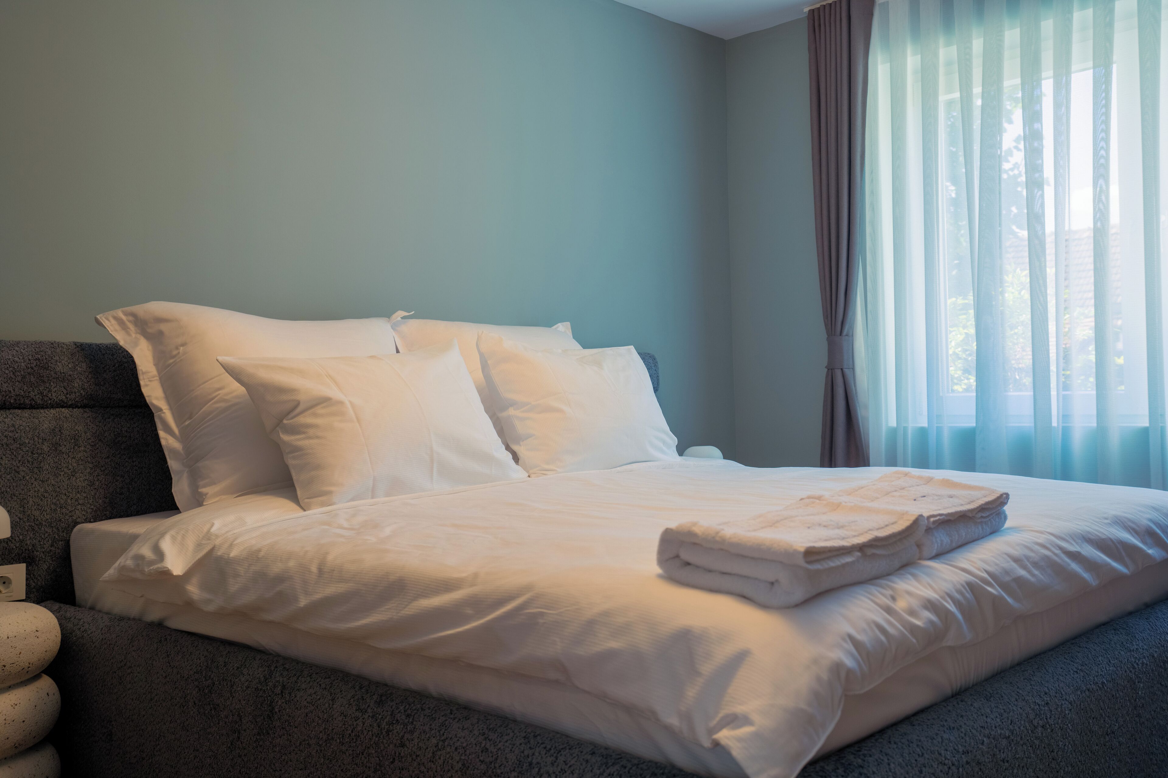 A neatly made bed with white linens and a folded blanket, a gray headboard, and a window with sheer curtains.