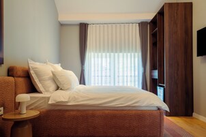 Un dormitorio moderno con una cama grande, una mesita de noche de madera, un televisor montado en la pared y una ventana con cortinas.