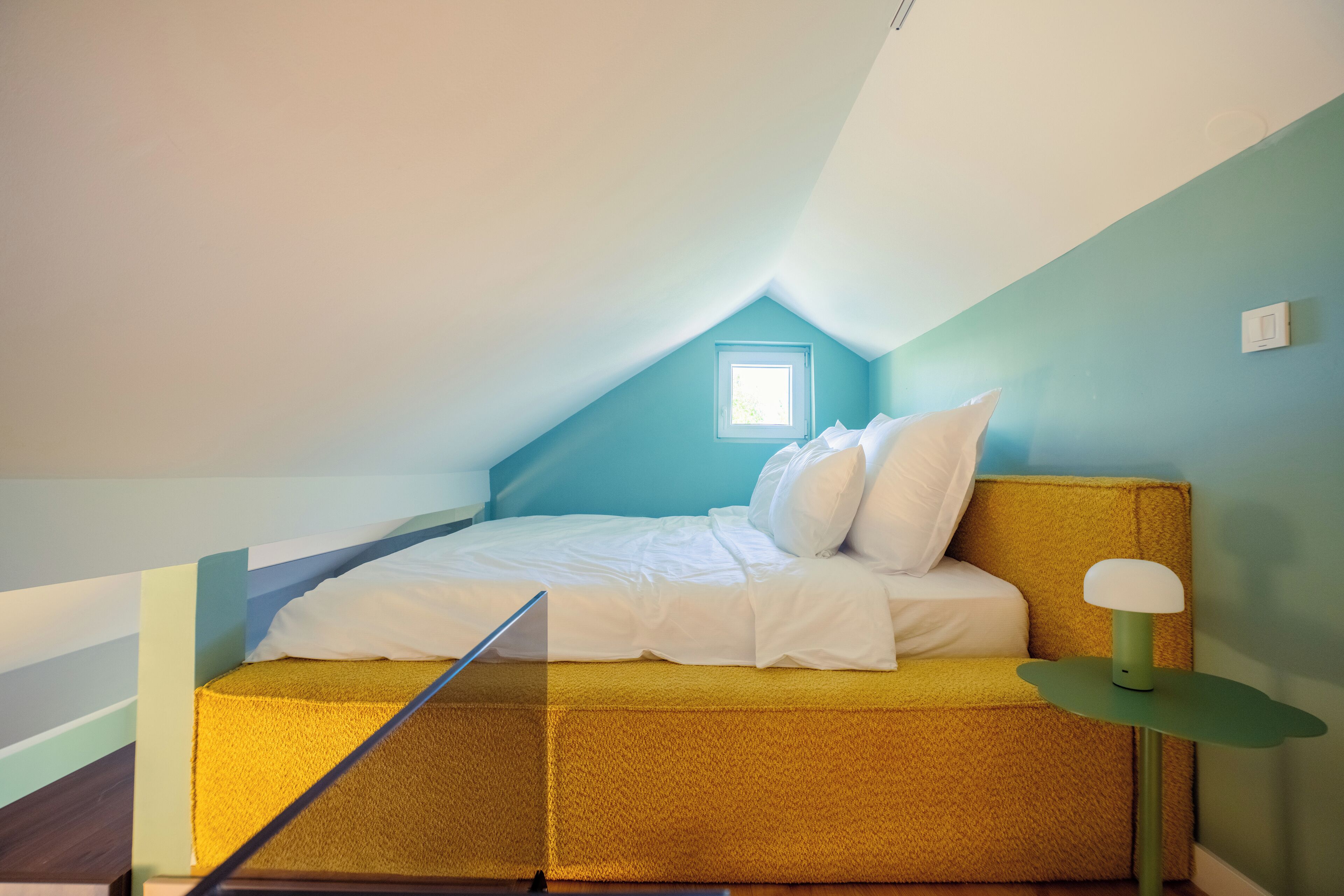 A small, cozy attic bedroom with a sloped ceiling, a single bed with white bedding, a green bedside table, and a small window providing natural light.