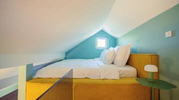 A small, cozy attic bedroom with a sloped ceiling, a single bed with white bedding, a green bedside table, and a small window providing natural light.