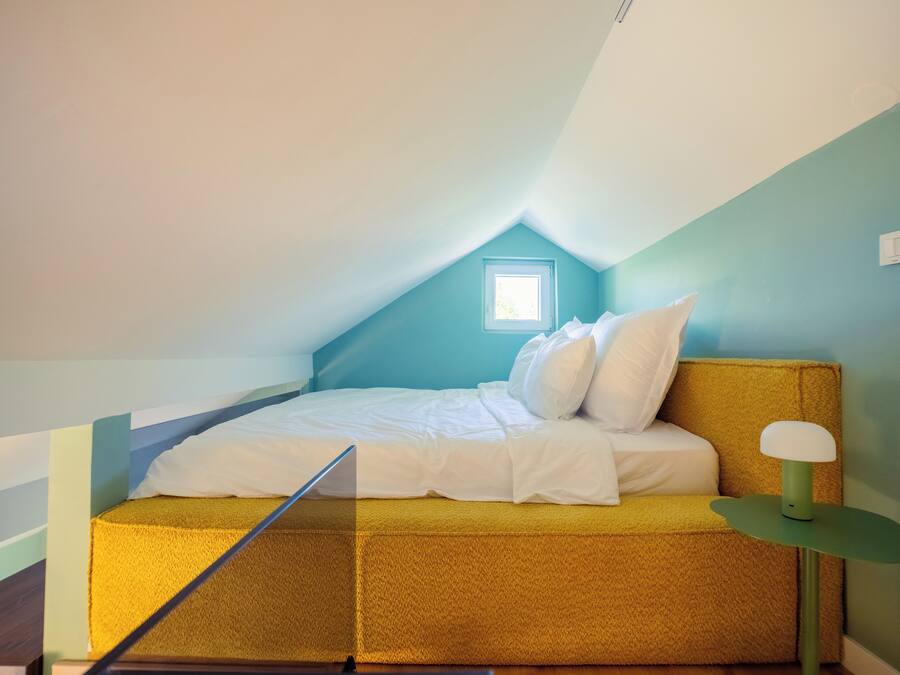 A small, cozy attic bedroom with a sloped ceiling, a single bed with white bedding, a green bedside table, and a small window providing natural light.