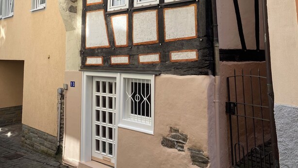 Ein traditionelles Fachwerkhäuschen mit mehreren Fenstern, einer Tür mit Metallgitter und einer gepflasterten Straße.