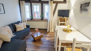 Ein gemütliches Wohnzimmer mit einem dunkelblauen Sofa, einem Holztisch und einem weißen Esstisch mit Stühlen. Zwei Fenster mit Vorhängen, ein Heizkörper und ein Fernseher auf einem Holzboden sind ebenfalls zu sehen.