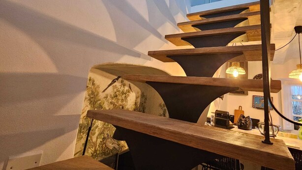 A modern interior with a wooden staircase leading to a mezzanine, a bar area with stools, and a view of the outside.