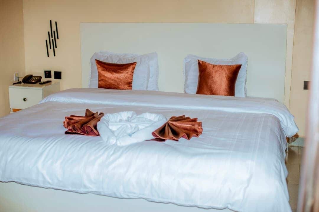 Una cama bien hecha con almohadas blancas y cobrizas, una toalla doblada y una mesita de noche con un teléfono.
