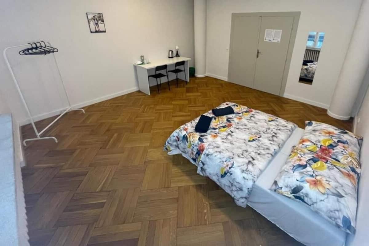 Ein Zimmer mit Holzboden im Fischgrätmuster, einem Bett mit floralem Überwurf, einem Schreibtisch mit Stühlen und einem Kleiderschrank.