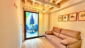 A room with a brown sofa, a wooden floor, a ceiling with exposed beams, a sliding glass door leading to an outdoor area with a table and chairs, and framed pictures on the wall.