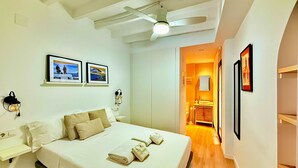 A hotel room with a bed, a ceiling fan, a nightstand, a wall-mounted lamp, and framed pictures on the wall.