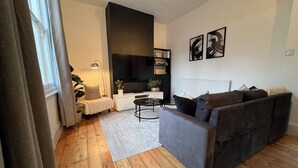 Ein modernes Wohnzimmer mit einem grauen Sofa, einem weißen Beistelltisch und einem schwarzen Couchtisch. Ein großes Fenster mit Vorhängen, eine Topfpflanze und ein Flachbildfernseher an der Wand.