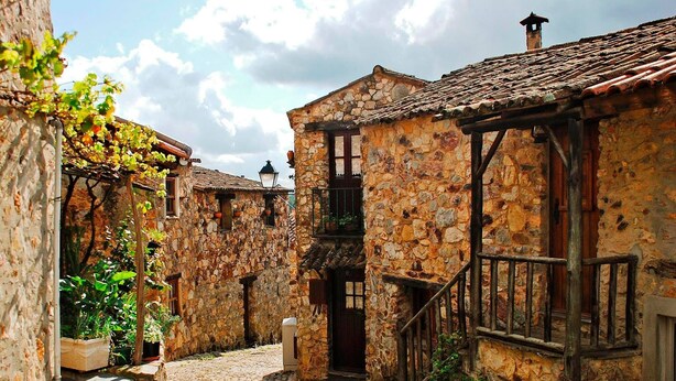 A narrow cobblestone street flanked by stone buildings with wooden balconies and a staircase leading to a door.