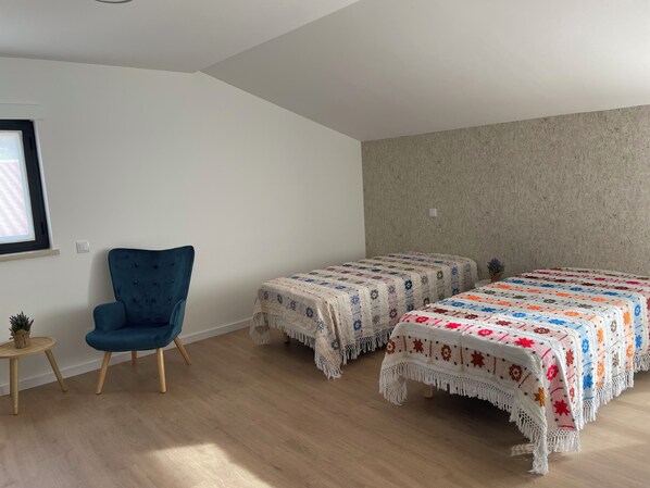 Quarto com duas camas, uma cadeira azul, uma mesa pequena e uma janela.