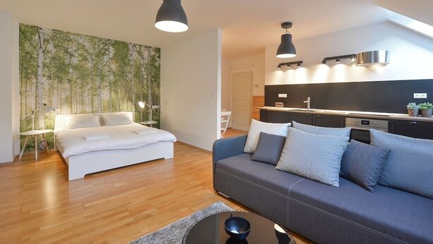 A modern bedroom with a bed, a sofa, and a dining area with a kitchen in the background.