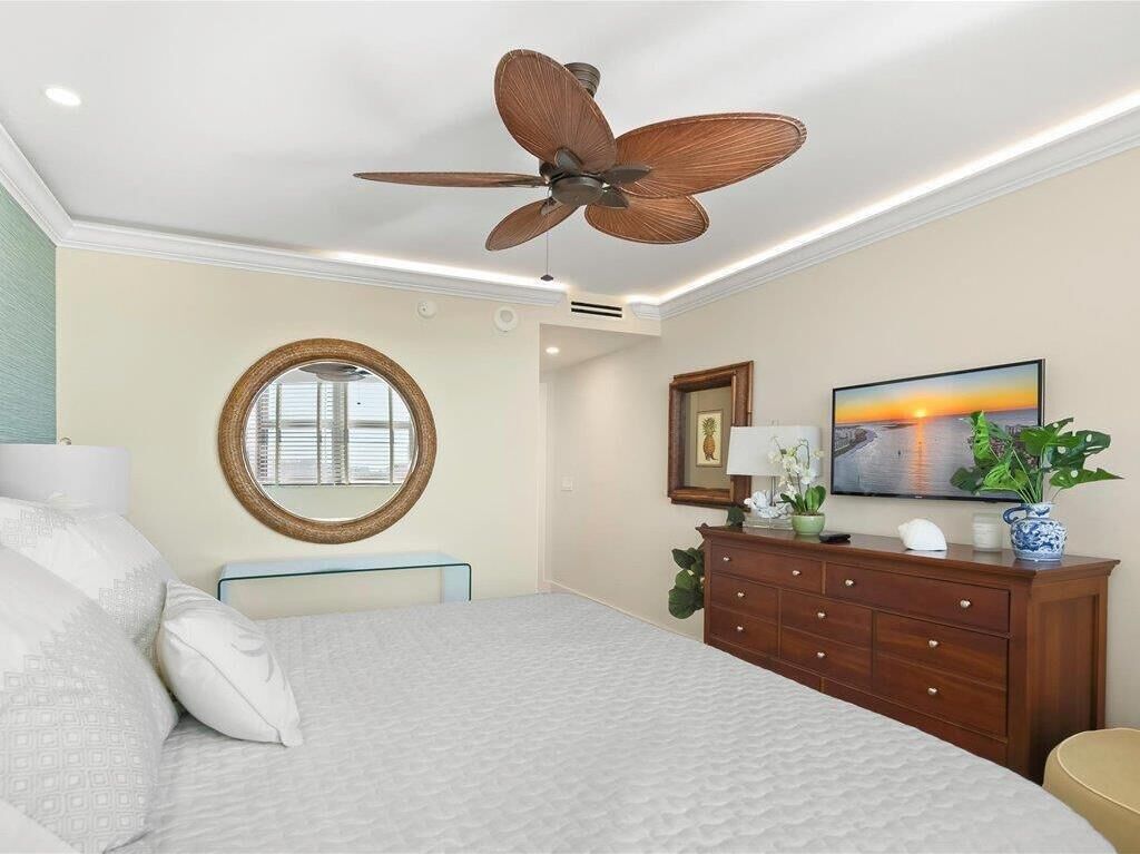 A bedroom with a bed, a wooden dresser, a round mirror, a ceiling fan, and a painting on the wall.
