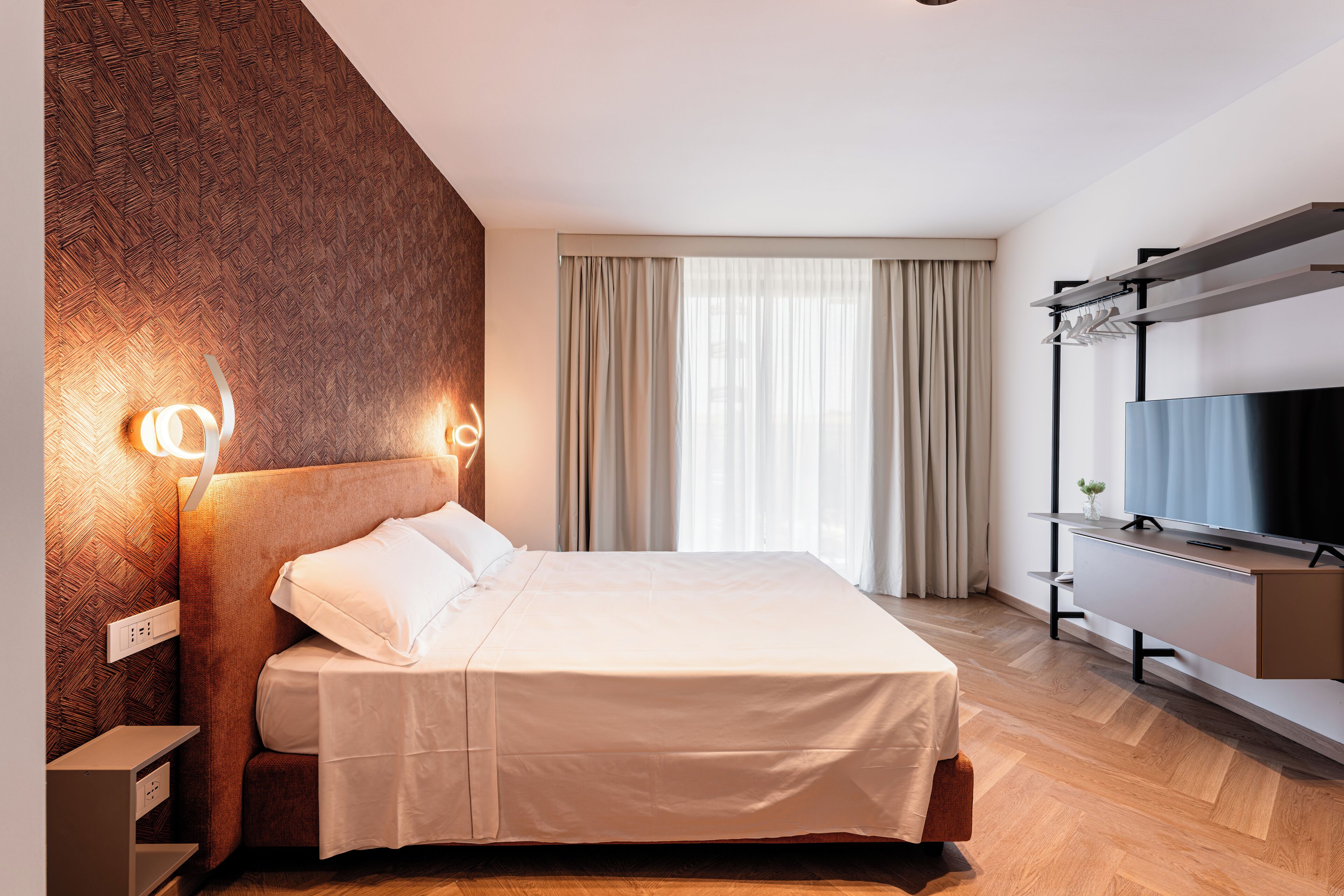 Une chambre d’hôtel moderne avec un grand lit, un lambris en bois, un téléviseur posé sur un meuble et une grande fenêtre avec des rideaux.