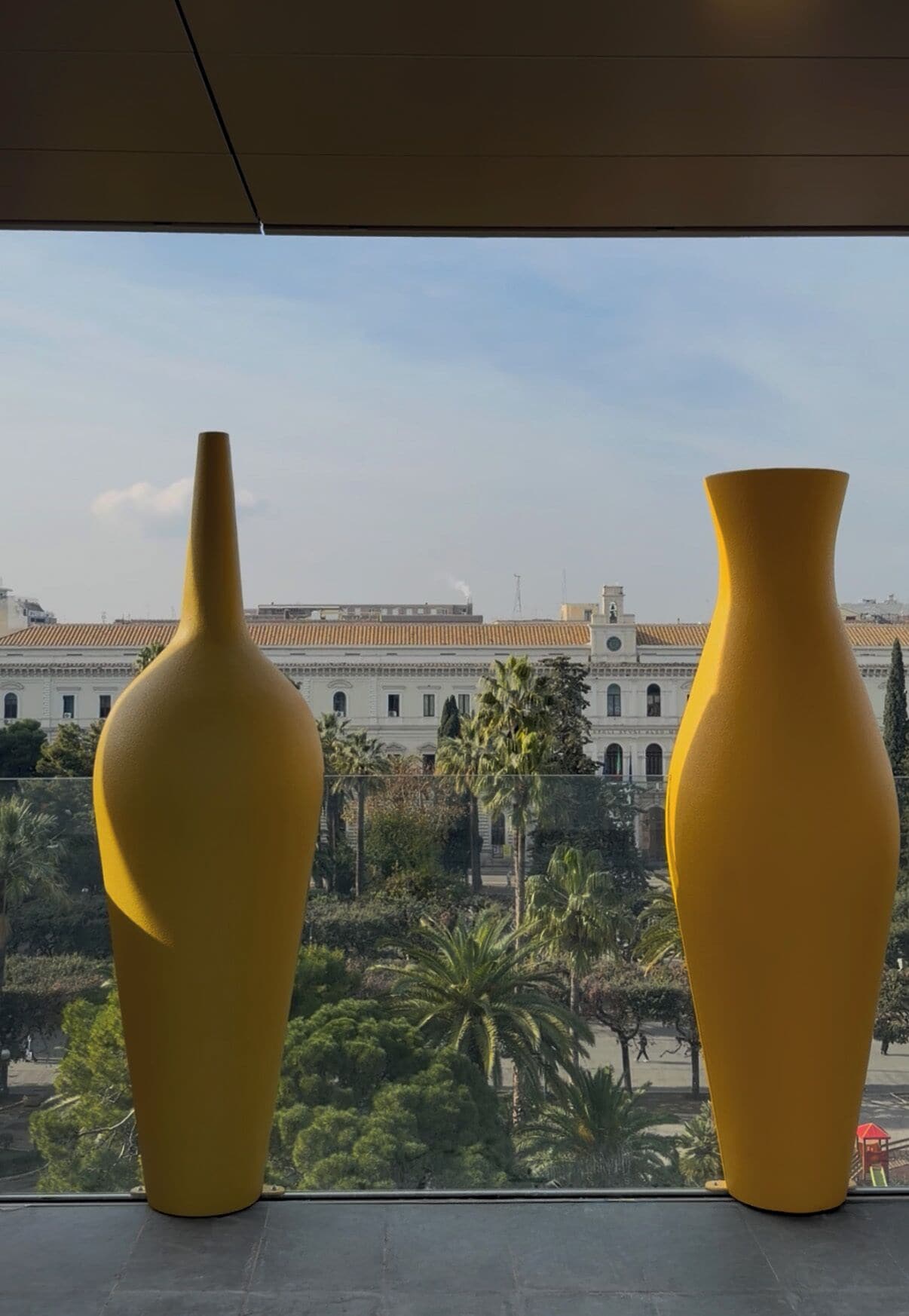 Dos jarrones amarillos en un alféizar con vistas a un edificio y palmeras.
