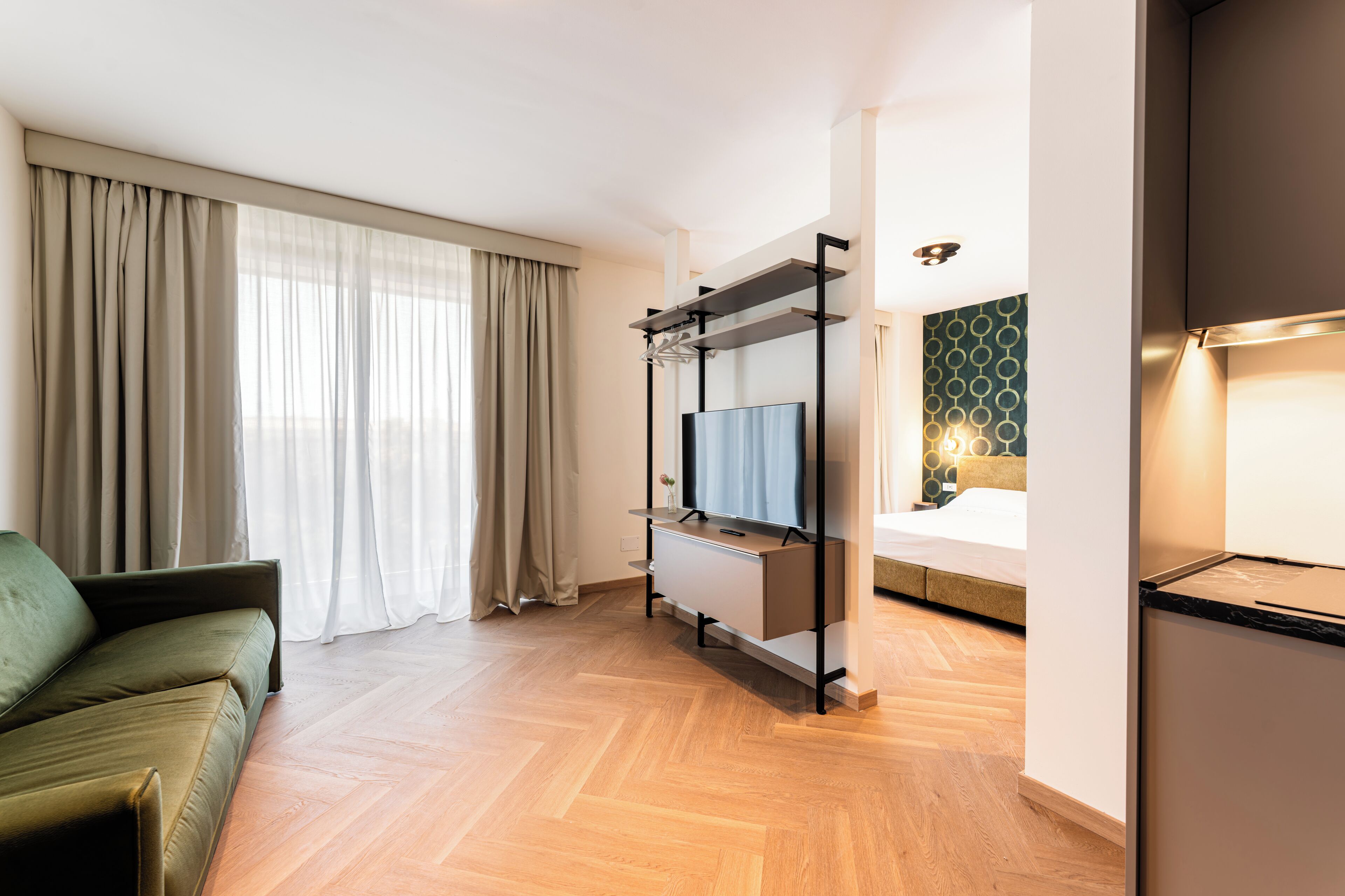 Een moderne hotelkamer met een bank, een televisie op een standaard, een bed met een hoofdeinde met een groen patroon, en een houten vloer.