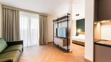 Una habitación de hotel moderna con sofá, televisor sobre un soporte, cama con cabecera estampada en verde y suelo de madera.