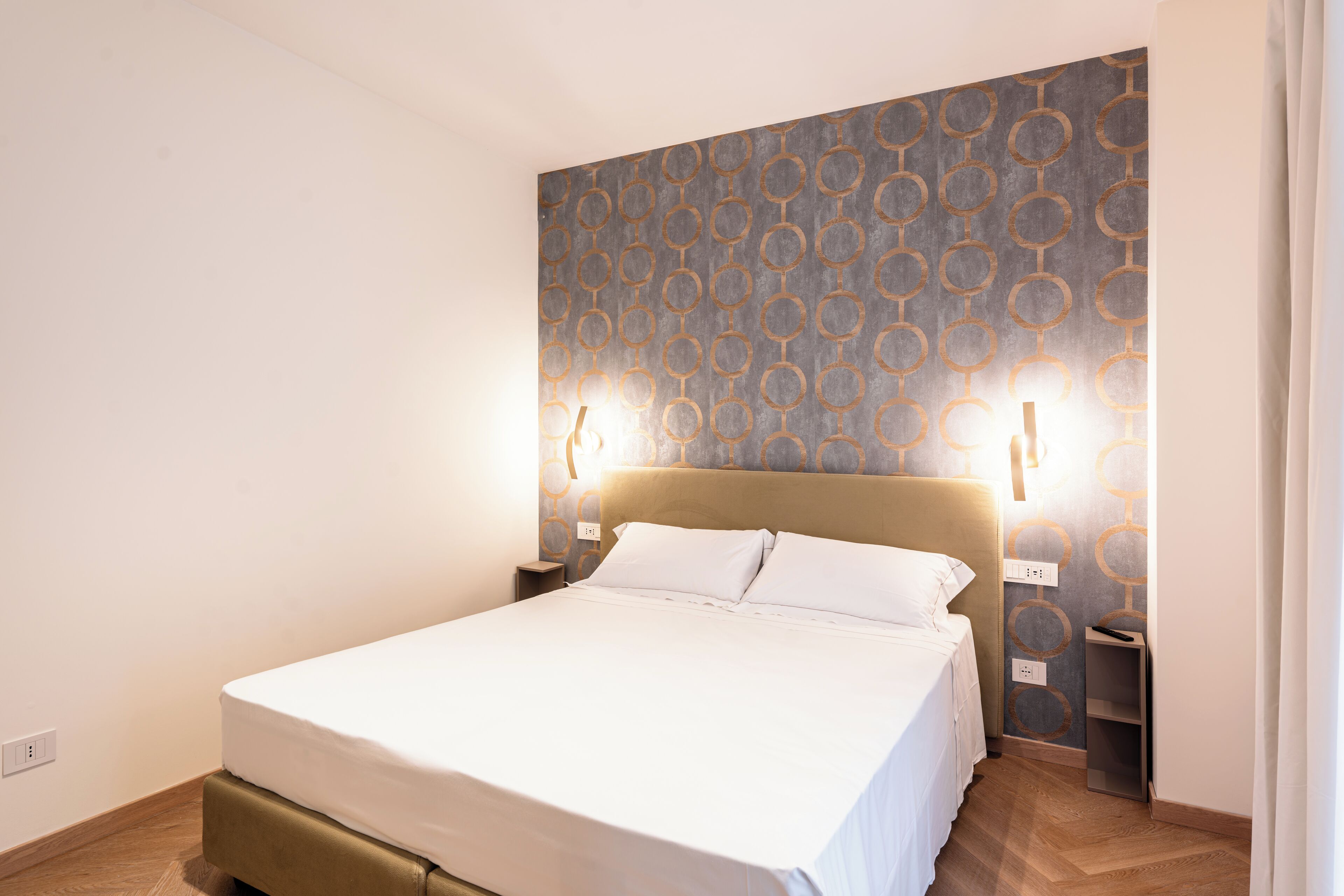 A neatly made bed in a hotel room with a patterned wall and two bedside lamps.