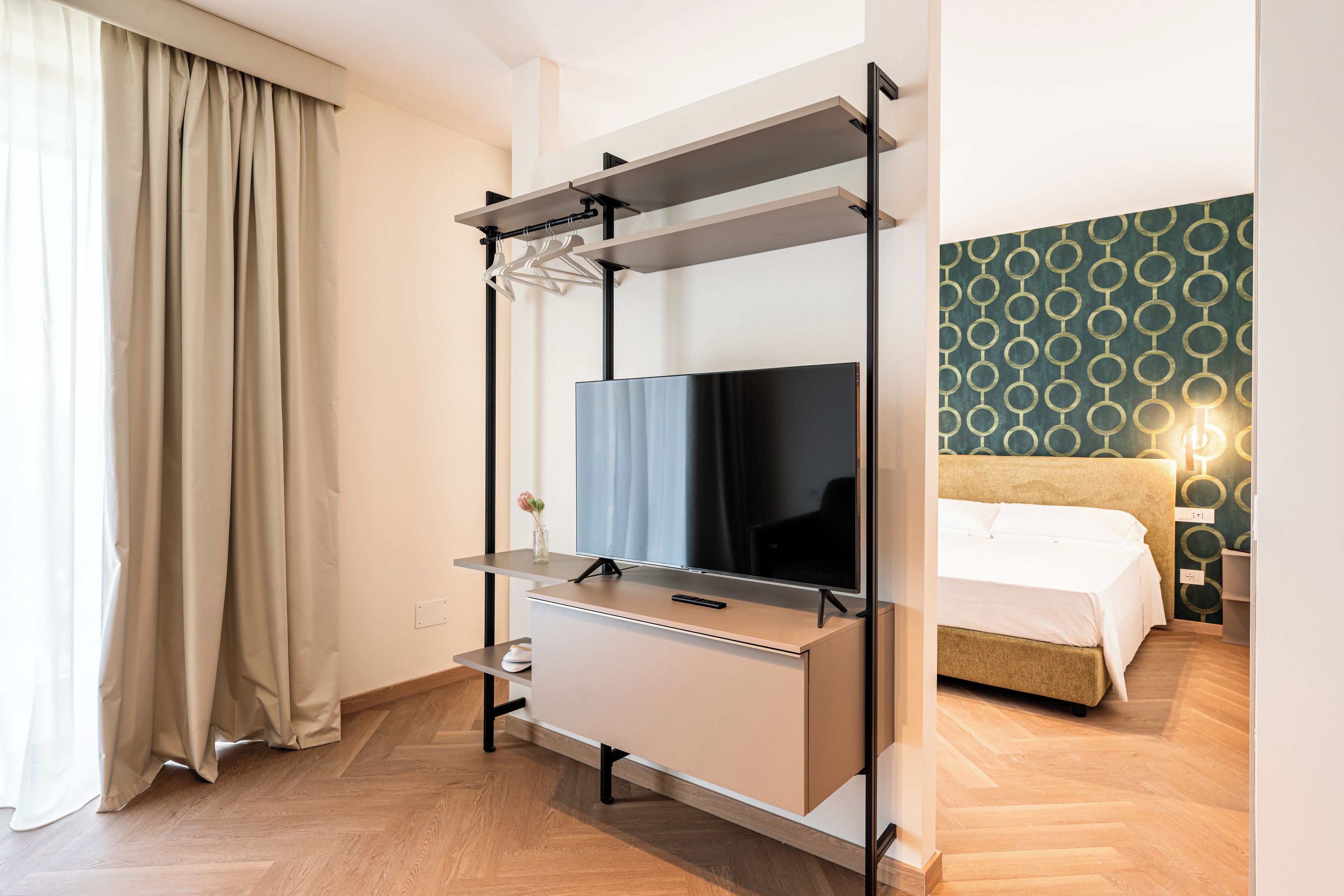 Habitación de hotel moderna con una televisión de pantalla plana sobre un soporte, una cama con cabecera y un papel tapiz estampado.