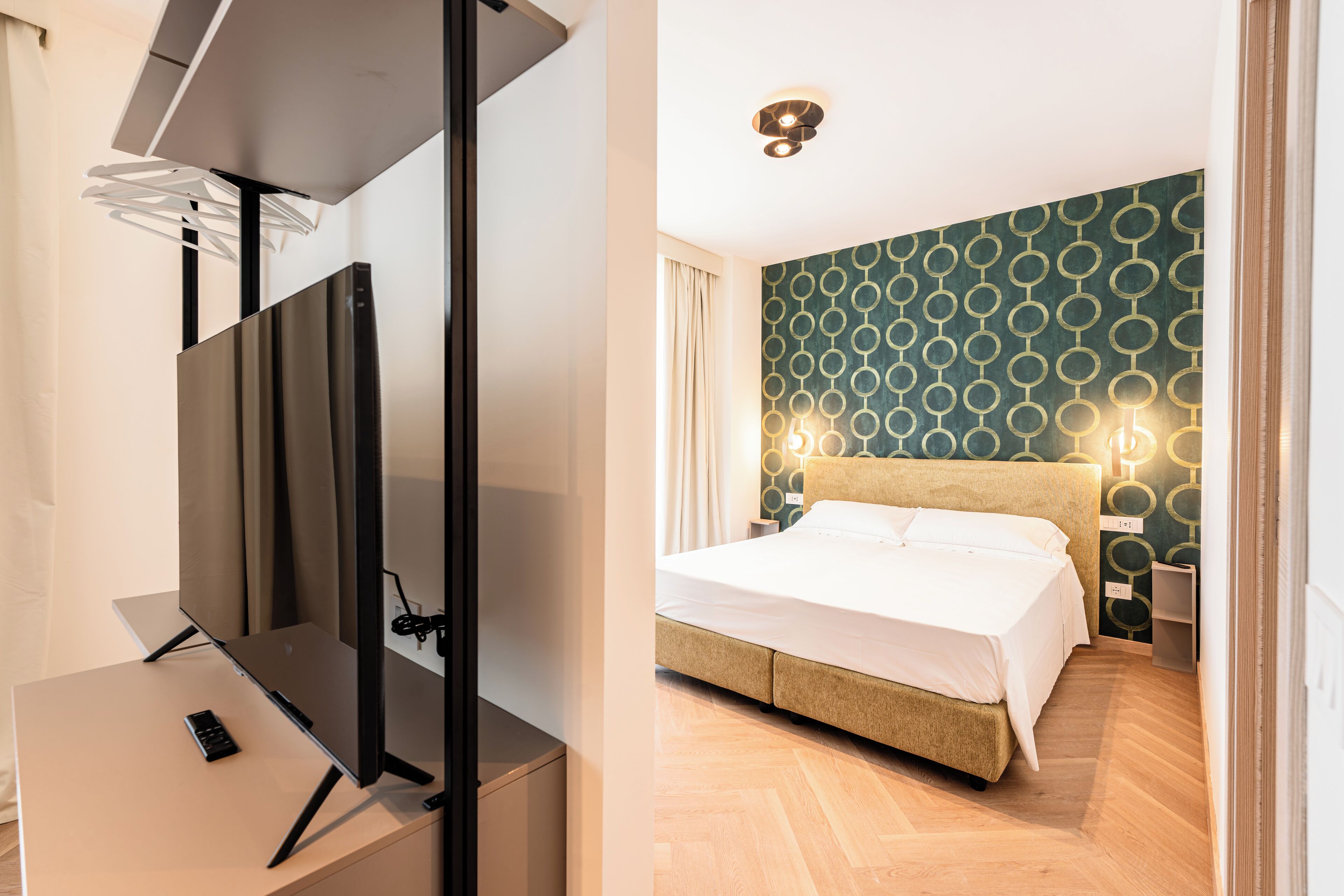 A modern hotel room with a large flat-screen TV, a bed with a headboard, and a patterned wall.