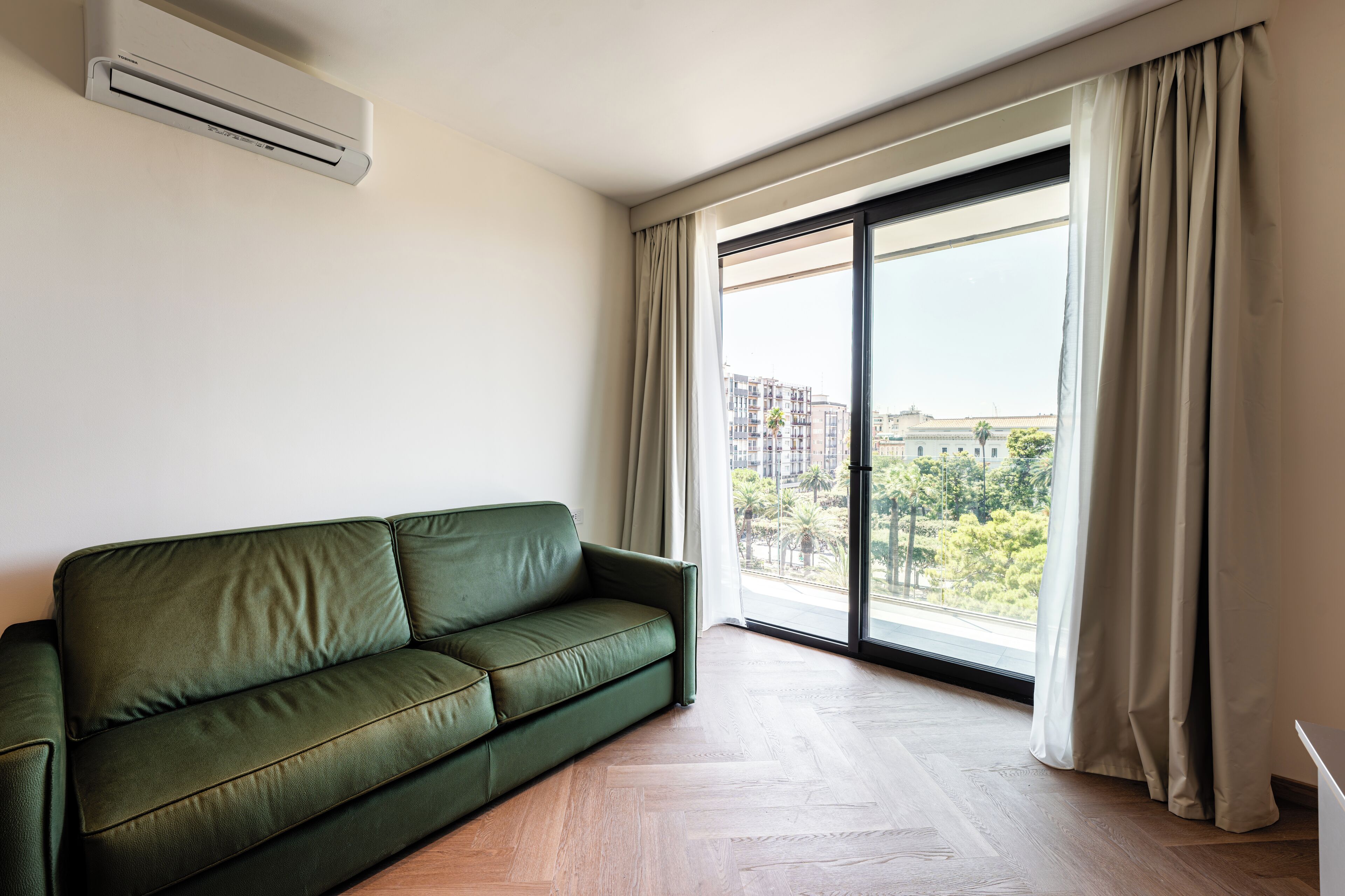 A green sofa in a room with a large window, a ceiling-mounted air conditioner, and a view of buildings and trees outside.