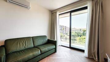 A green sofa in a room with a large window, a ceiling-mounted air conditioner, and a view of buildings and trees outside.