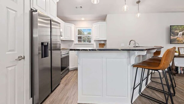 A modern kitchen with white cabinetry, a stainless steel refrigerator, a built-in island with bar stools, pendant lighting, and a window with blinds. - Quiet Razorback Escape - Easy Drive to Downtown & Stadium (Fayetteville)
