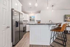 A modern kitchen with white cabinetry, a stainless steel refrigerator, a built-in island with bar stools, pendant lighting, and a window with blinds. - Quiet Razorback Escape - Easy Drive to Downtown & Stadium (Fayetteville)