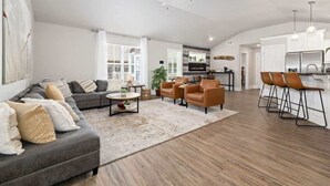 A spacious living room with a gray sofa, a coffee table, and a fireplace. There is a kitchen area with white cabinets and a dining area with bar stools. - Quiet Razorback Escape - Easy Drive to Downtown & Stadium (Fayetteville)