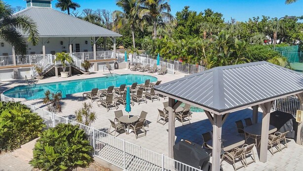 A pool area with lounge chairs and a covered seating area, surrounded by palm trees and a building with a grey roof.