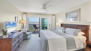 A hotel room with a bed, a TV, a desk with a lamp, a chair, and a balcony with a view of the beach. - Boca Grande North - 08: 2BR condo with water views, pool, and hot tub in Boca Grande, FL. (Boca Grande)