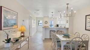 A spacious kitchen with a central island, a dining area with a glass table, and a hallway leading to a door. - Boca Grande North - 08: 2BR condo with water views, pool, and hot tub in Boca Grande, FL. (Boca Grande)