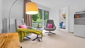 Ein modernes Wohnzimmer mit Sofa, Sessel und Couchtisch. Ein Bücherregal und ein Fenster mit Blick auf Grünpflanzen.