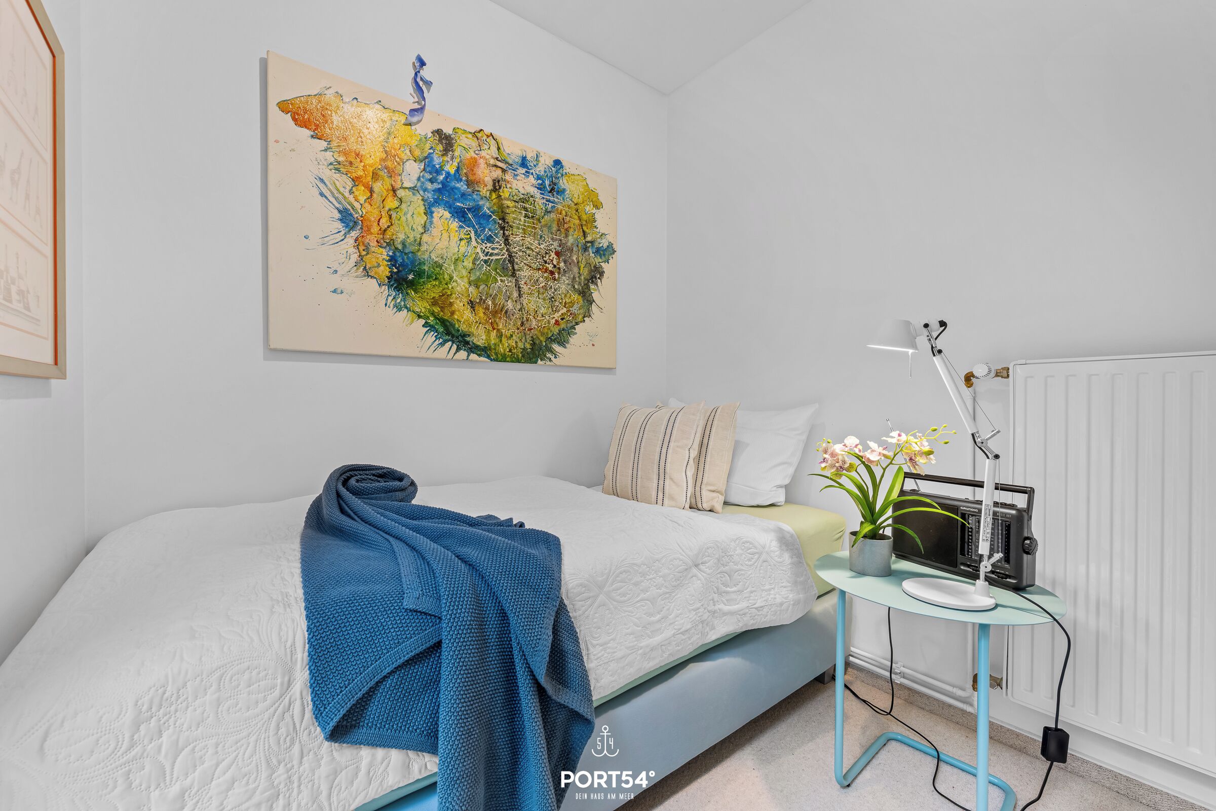 Quarto com cama, mesa de cabeceira com abajur, radiador e um quadro abstrato colorido na parede.