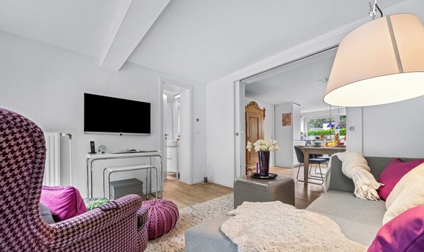 Una sala de estar moderna con un sofá, un sillón y un televisor. En el fondo, se observa un comedor con una mesa y sillas.