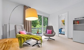 Un salon moderne avec un canapé, un fauteuil et une table basse. On y trouve également une bibliothèque et une fenêtre donnant sur un paysage verdoyant.