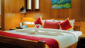A hotel room with a wooden bed, red and white bedding, a heart-shaped towel decoration, a painting of a mountain landscape, and a nightstand with a kettle and bottles. - Anvitha Mansions (Chikkamagaluru)