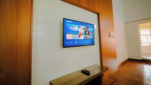 A flat-screen TV mounted on a wooden wall with a remote control on a glass-topped table below. - Anvitha Mansions (Chikkamagaluru)