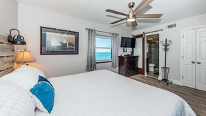 Un dormitorio con cama, ventilador de techo, televisor, ventana con cortinas, cómoda y perchero.