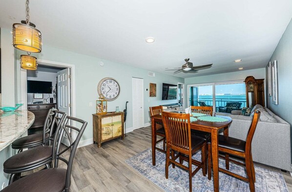 A dining area with a wooden table and chairs, a clock on the wall, and a view of the ocean through large windows.