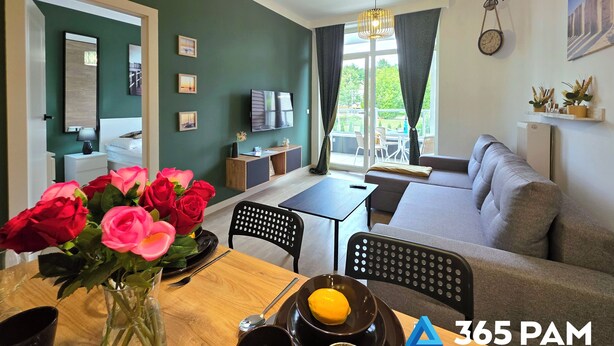 A modern living room with a sofa, dining table, and a TV. There are flowers on the table and a clock on the wall. - Apartament Ku Morzu 27 - 365PAM (Sianożęty)