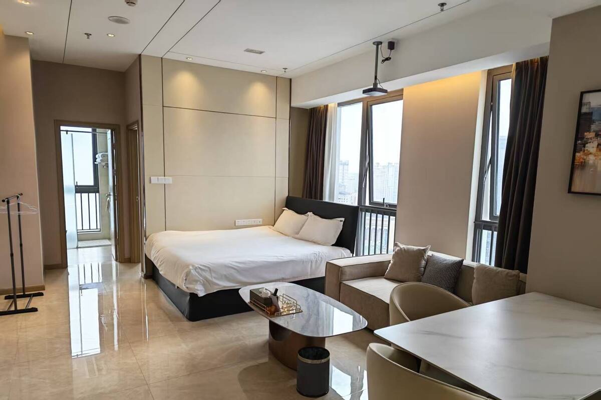 A modern hotel room with a bed, a sofa, a dining table, and a trash bin.