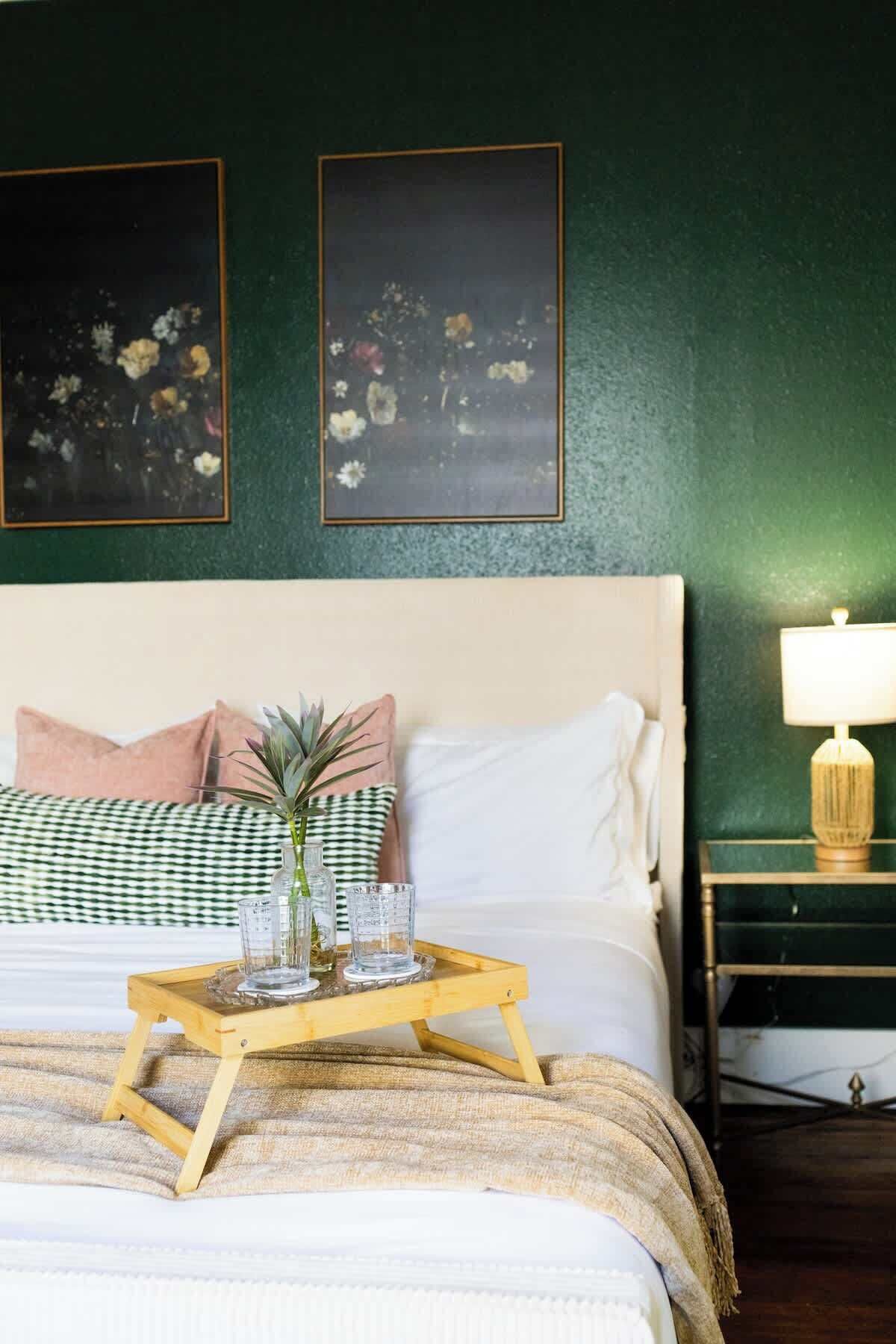 Un lit bien fait, avec des oreillers, une table de chevet avec une lampe, et un plateau avec des verres et une plante.